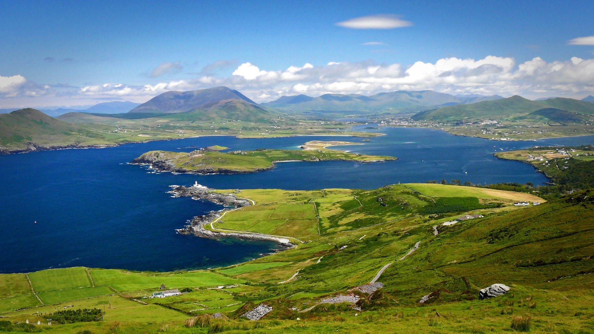 Aerial view of the breathtaking Ring of Kerry, a scenic drive in County Kerry, Ireland, showcasing the rugged coastline, rolling green hills, and sparkling lakes, with the majestic MacGillycuddy's Reeks mountains rising in the distance.