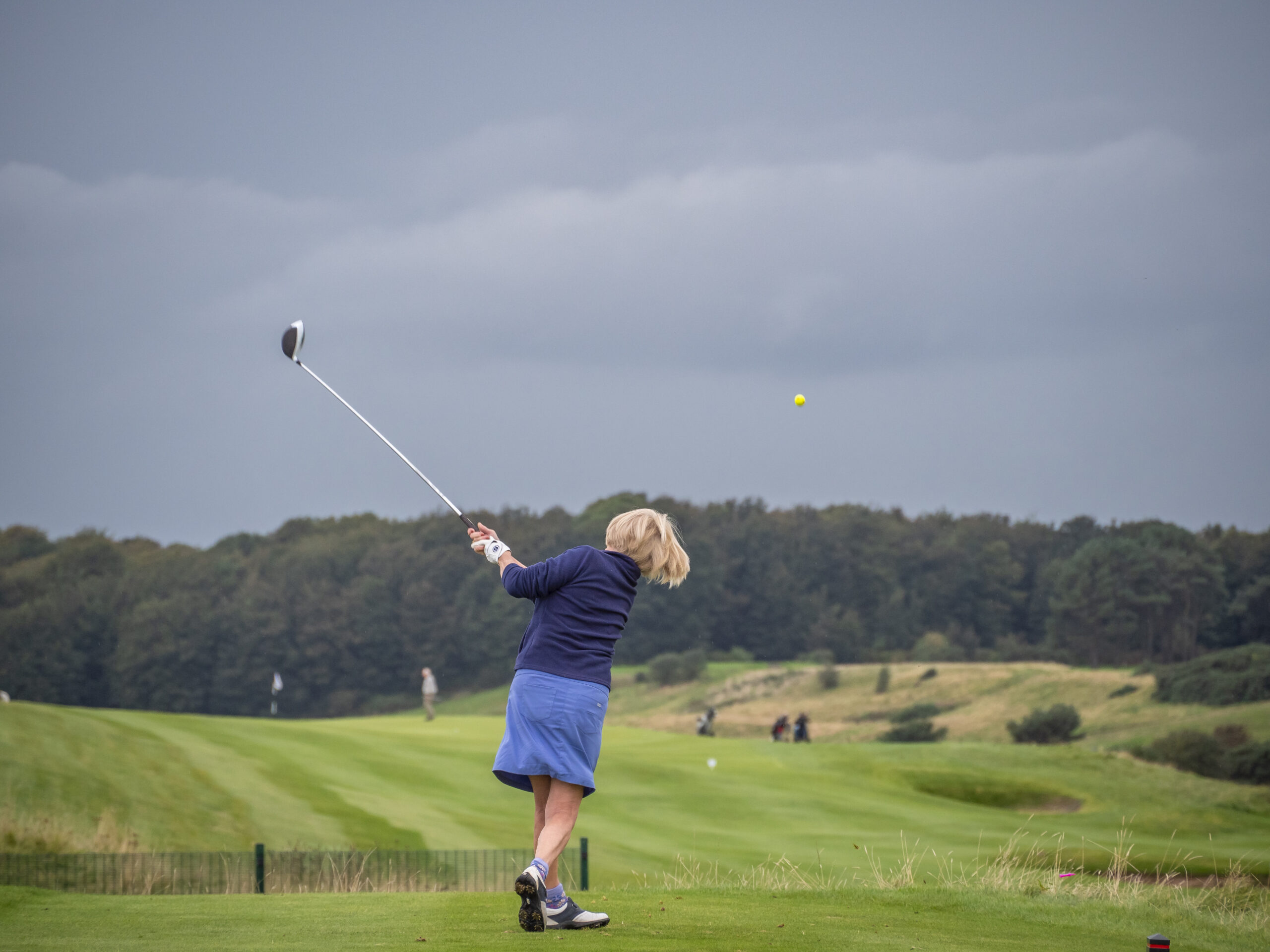 A lady golfer taking a powerful swing at Lundin Links Golf Club, with the stunning backdrop of the Firth of Forth and the lush, green fairways of the links-style course.