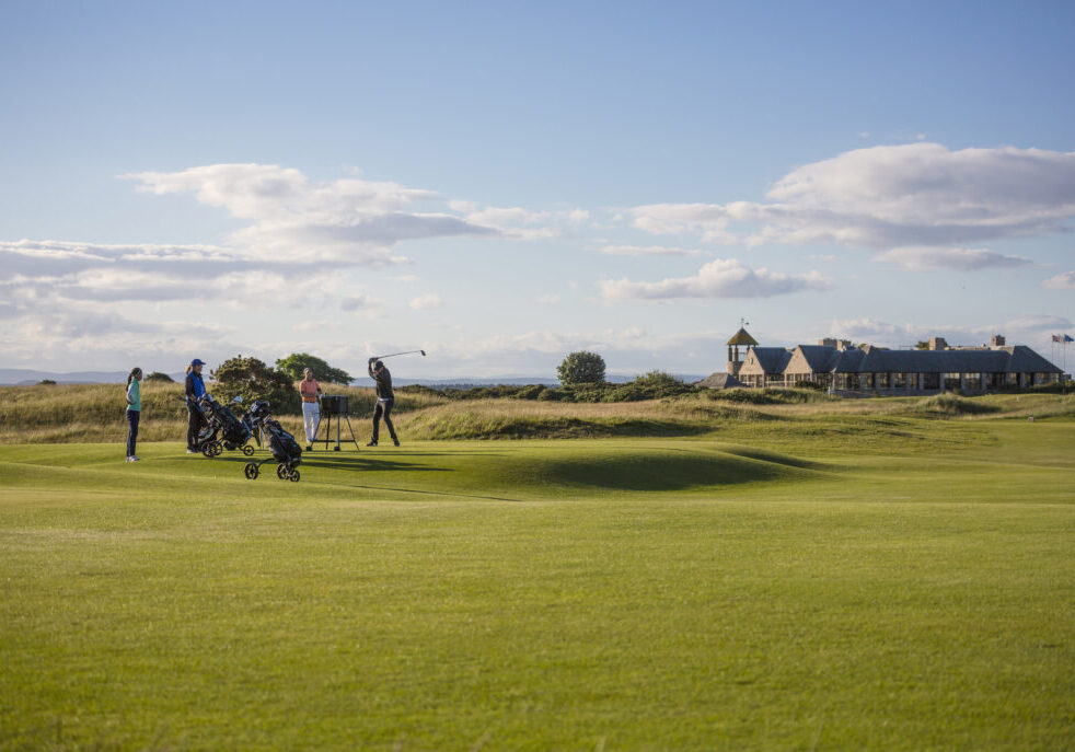 St. Andrews Old Course: The historic and iconic golf course with its rolling fairways, famous Swilcan Bridge, and the clubhouse in the distance, set against a backdrop of a clear blue sky.